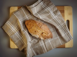 Freshly baked, hot country bread with flour, freshly from the oven on a brown linen towel with white squares, a wooden board. Appetizing photography. It resembles an American football ball in shape.