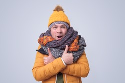 Handsome caucasian shocked funny man in several hats and scarfs showing thumb up gesture on colored background. Winter fashion.