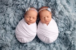 Tiny newborn twin girls in white cocoons on a blue background. A newborn twin sleeps next to his sister. Newborn twin girls in white headbands with a white and blue flowers. 