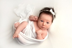 Top view of a newborn baby girl lying in a white cocoon on a white bed with a white bandage. Beautiful portrait of a little girl with wide open eyes 7 days, one week. Photography in macro studio.