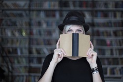 Portrait of woman in black hat with opened book, face half-covered, white hair. Hipster student girl in a library