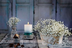 A bouquet of white gypsophila stands in an antique metal vase, in the background a glass vase on the old wood decorated with gray fabric. Background is light blue, old wooden door. Candle burns.