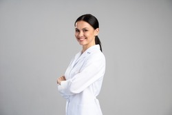 Side view of the smiling female doctor in lab coat with arms crossed looking away and posing against grey background. Medicine concept 
