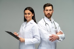 Woman doctor holding clipboard and posing with her male colleague with stethoscope with happy faces while standing isolated on grey background 