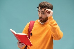 Shocking facts. Astonished smart bearded student man reading book with surprised expression, amazed by story, being shocked by interesting information. Indoor studio shot isolated on blue background 