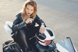 Rebel freedom concept. Young beautiful biker woman sitting with bent legs on a motorbike in a fashionable pose. Woman wearing leather clothes sitting on a white motorcycle holding arm on helmet