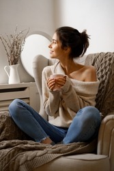 Smiling lady in smart trendy wear is sitting on armchair with a cup of tea. Smiling, sitting in relaxing atmosphere indoors at home, nice design.