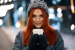 Girl drinking hot coffee while walking in the evening city market decorated with holiday lights in the evening. Feeling happy in big city. Spending winter vacations