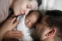 Top view of the pretty caucasian couple with dark hair laying with their beautiful newborn baby at the bed and enjoying time with him. Cute child sleeping between of his parents