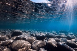 Underwater view with stones bottom, sun rays and reflection in sea water.