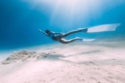 Freediver underwater glides with white fins. Attractive woman free diver in blue ocean with sun rays