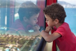 A cute boy in a red t-shirt thought looking out the window. The child and his reflection in the glass.