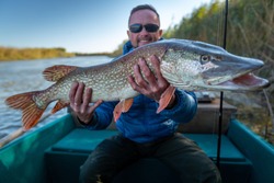Angler with pike fish. Amateur fisherman holds trophy pike (Esox lucius) and sits in the boat with river on the background