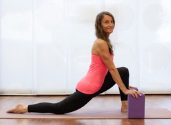 Middle aged beautiful yogi practicing dragon pose with hands on purple blocks. Smiling woman on pink tank top doing yin yoga over white background at studio. Active, healthy lifestyle concepts