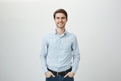 Confidence and business concept. Portrait of charming successful young entrepreneur in blue-collar shirt, smiling broadly with self-assured expression while holding hands in pockets over gray wall