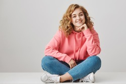 Girl is always happy to hear interesting stories. Curious cheerful woman with curly hair in stylish outfit sitting on floor with crossed legs, leaning on hand, smiling, taking part in conversation