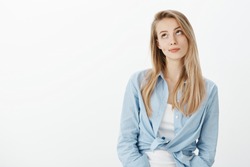 Portrait of unimpressed beautiful european woman with blond hair, smirking and looking upwards with indifferent expression, standing in stylish outfit over white background casually