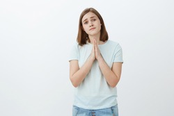 Girl really needs help asking it with cute angel face. Tender and attractive young girlfriend in t-shirt and jeans tilting head smiling gentle, holding hands in pray while begging favor or apologizing