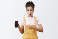 Female friend showing girlfriend strange message she received, holding smartphone and pointing at screen with index finger, folding lips from interest and surprise, standing shocked and impressed