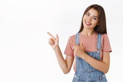 Lovely, alluring cheerful beautiful woman in dungarees, t-shirt, tilt head silly and smiling as promote company product, pointing upper left corner, give advice to check out cool advertisement