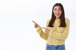 Excited and surprised young happy woman in yellow sweater, drop jaw gasping in awe and astonished pointing looking left side blank space for your promo, standing white background fascinated