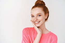 Beauty and skincare. Smiling girl with ginger hair combed in messy bun, touching perfect skin and smiling, standing without makeup on white background.