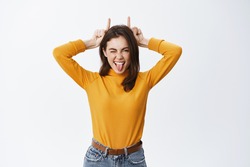 She is a rebel. Beautiful young woman showing tongue and finger devil horns on head, being stubborn, winking sassy at camera, standing against white background.