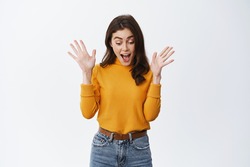 Excited girl screaming of fantastic news, gasping and raising hands up, looking down at amazing thing, checking out promo offer, standing against white background.