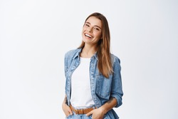 Happy carefree woman with joyful expression, smiling while standing in relaxed pose against white background, holding hands in jeans pockets