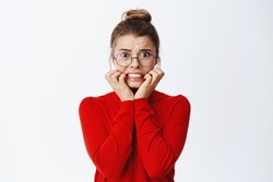 Image of scared young female employee in glasses panicking, biting finger nails from fear and anxiety, trembling of something scary, standing over white background