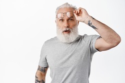 Surprised mature guy with long beard, take-off glasses and look in awe, staring at something amazing, standing in grey t-shirt against white background