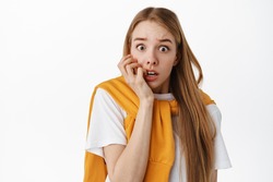 Close up of shocked and scared blond woman, biting fingers and gasping, staring startled and frightened, afraid of something scary, standing over white background