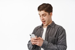 Close up shot of surprised young man staring at phone in awe, checking out awesome news on smartphone, receive notification app, standing over white background