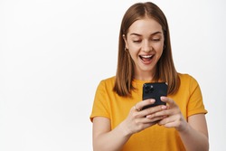 Portrait of girl looks surprised and excited at smartphone screen, receive pleasant notification, watching video on mobile phone, cellular internet, white background