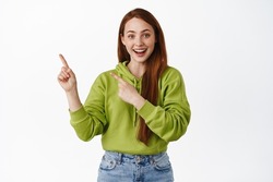 Fantastic promo ahead. Smiling redhead teen girl shows information, pointing left at advertisement and looking happy, standing over white background