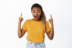 Surprised and excited Black woman pointing fingers up, gasping in awe, amazed by price sale, standing in yellow t-shirt over white background