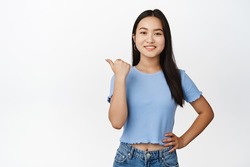 Smiling beautiful asian girl pointing left, looking confident and happy, showing way, sale banner, standing in tshirt over white background