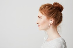Profile portrait of young beautiful tender redhead girl with bun in headphones smiling over white background.
