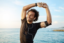 Portrait of a smiling afro-american sports man stretching his muscular arms before workout by the sea, using music app on his smartphone. Dark-skinned athlete warming up before running.