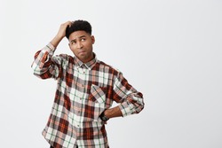 Portrait of young confused dark-skinned handsome man with afro hairstyle in checkered shirt holding head with hand, looking aside with satisfied expression, don't know what to do with debts
