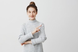 Pretty brunette woman posing on camera with tricky look and gesturing to side. Female designer presenting lovely product with index finger. Gesture concept