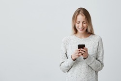 Blonde smiling young woman demonstrating white teeth using cell phone, messaging, being happy to text with her boyfriend, looking at screen of smartphone. Modern technologies and communication