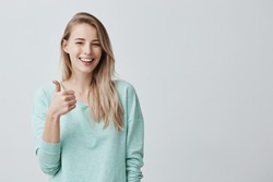 Happy young caucasian female wearing blue long sleeved shirt making thumb up sign and smiling cheerfully, showing her support and respect to someone. Body language. I like that. Good job.