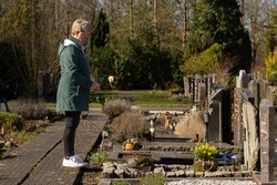 An elderly lady with a yellow rose stands at the grave of a loved one.