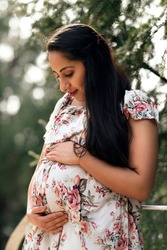 Portrait of pregnant woman with long black hair in park dressed in white dress with flowers, woman holds her baby belly, pregnant woman in stands in park, indian woman portrait