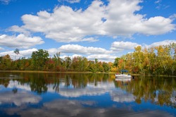 Photograph of a quiet and remote northwoods lake on a beautiful autumn day with clouds reflected in the still blue waters while a pontoon boat of sightseers slowly motors by.