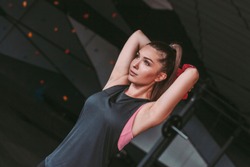 Portrait of beautiful sports woman lifting dumbbells from behind head while standing in dark gym. Athletic young girl in sports wear doing exercises for triceps with dumbbells. Muscle training in gym
