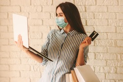 Young beautiful woman shopper in protective face mask hold laptop, credit card and shopping bags in hand. Happy girl in medical face mask shopping at online store. Coronavirus COVID-19, sale, discount