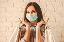 Beautiful girl shopper in medical face mask holding shopping bags in hands. Young happy woman customer in protective mask on face with paper bags. Girl shopaholic. Coronavirus COVID-19, sale, discount