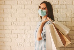 Beautiful girl in protective face mask holding shopping bags in hands. Young woman shopaholic in medical mask on face with paper bags. Female shopper. Safe shopping, coronavirus COVID-19, copy space